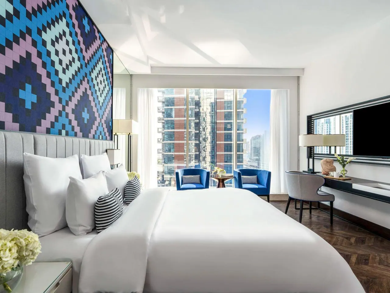 A bedroom with a large white bed, a patterned feature wall, blue armchairs by a floor-to-ceiling window showing high-rise towers, and a wall-mounted television beside a desk.