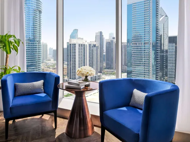 Two blue armchairs and a round side table with flowers and books sit in front of floor-to-ceiling windows overlooking a city of high-rise towers.
