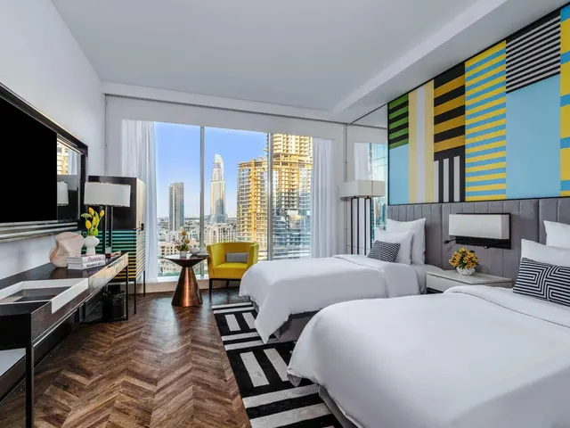 A bedroom with two single beds, a television, a yellow armchair and a floor-to-ceiling window looking out over city towers including the Burj Khalifa.