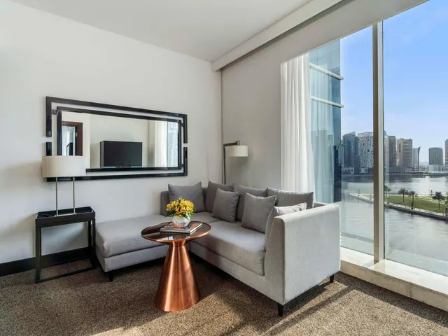 A living area with a grey sectional sofa, a copper side table with yellow flowers, a wall mirror and floor-to-ceiling windows overlooking a canal and city towers.
