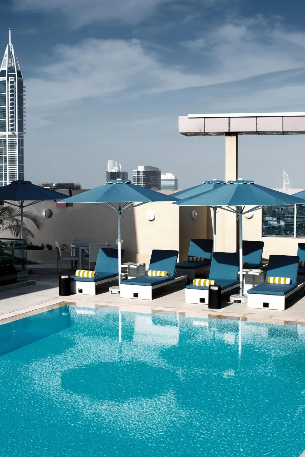 A rooftop swimming pool with blue sun loungers and umbrellas, with city towers visible in the background.