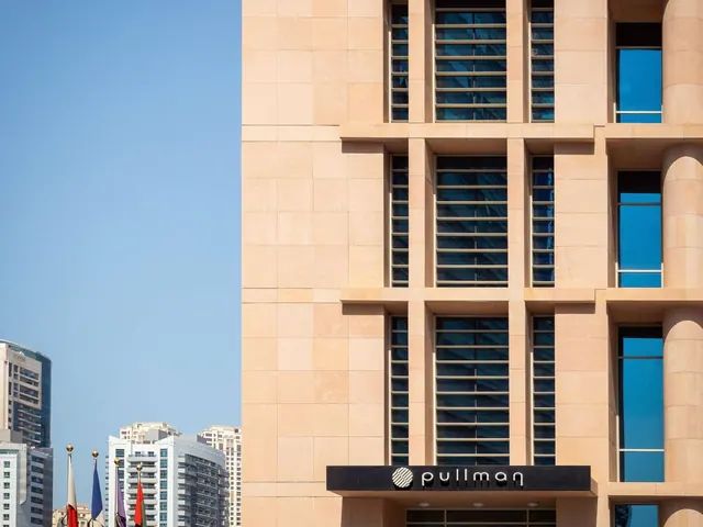 The exterior of a sandstone-coloured hotel building with a Pullman sign above the entrance, under a clear blue sky, with flags and towers visible at the left edge.