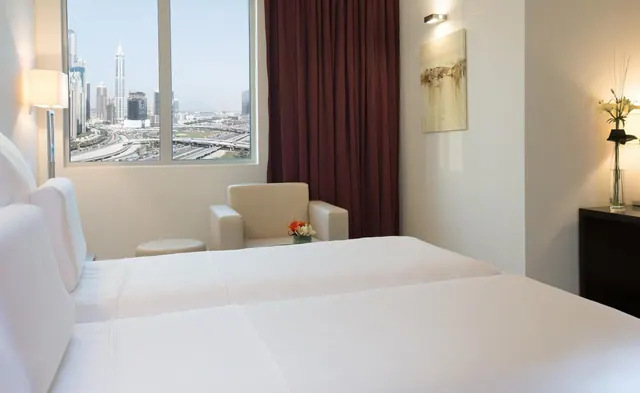 A bedroom with two white beds, an armchair and a small side table, and a window looking over a city skyline and a highway interchange.