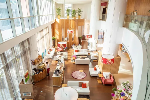Elevated view of a double-height hotel lobby with white sofas, armchairs, potted plants, red lampshades and a staff member at a counter on the left.