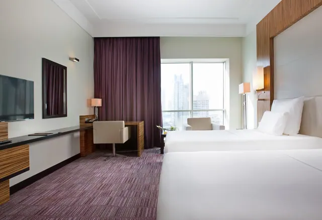 A hotel bedroom with two white beds, a wooden desk with a wall-mounted television, an armchair, and a window showing city skyscrapers.