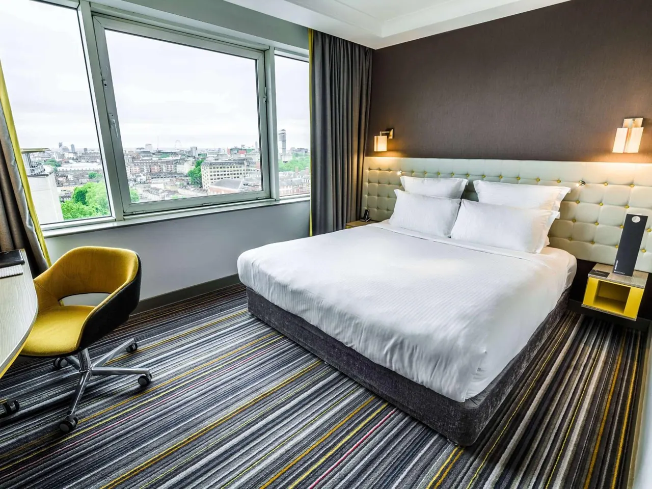 A king bed with white bedding stands beside a large window overlooking London rooftops, with a yellow desk chair at the left edge.