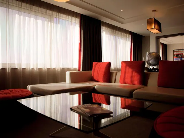 A living area with a beige sectional sofa, red cushions, a glass coffee table holding magazines, and sheer curtains over large windows.