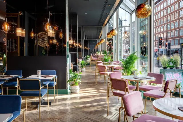 A restaurant dining room with blue and pink velvet chairs, round marble-topped tables, copper pendant lamps and tall windows onto a street.