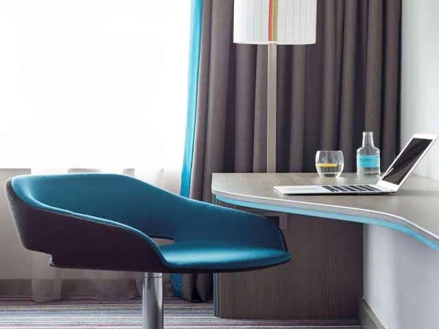 A blue swivel armchair sits beside a wooden desk holding an open laptop, a glass of water and a bottle of water, with grey curtains and a floor lamp behind.