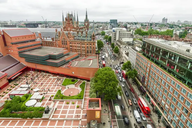 Aerial view over a busy road with traffic, showing a Gothic red-brick hotel, a large red-brick building and a paved plaza with trees.
