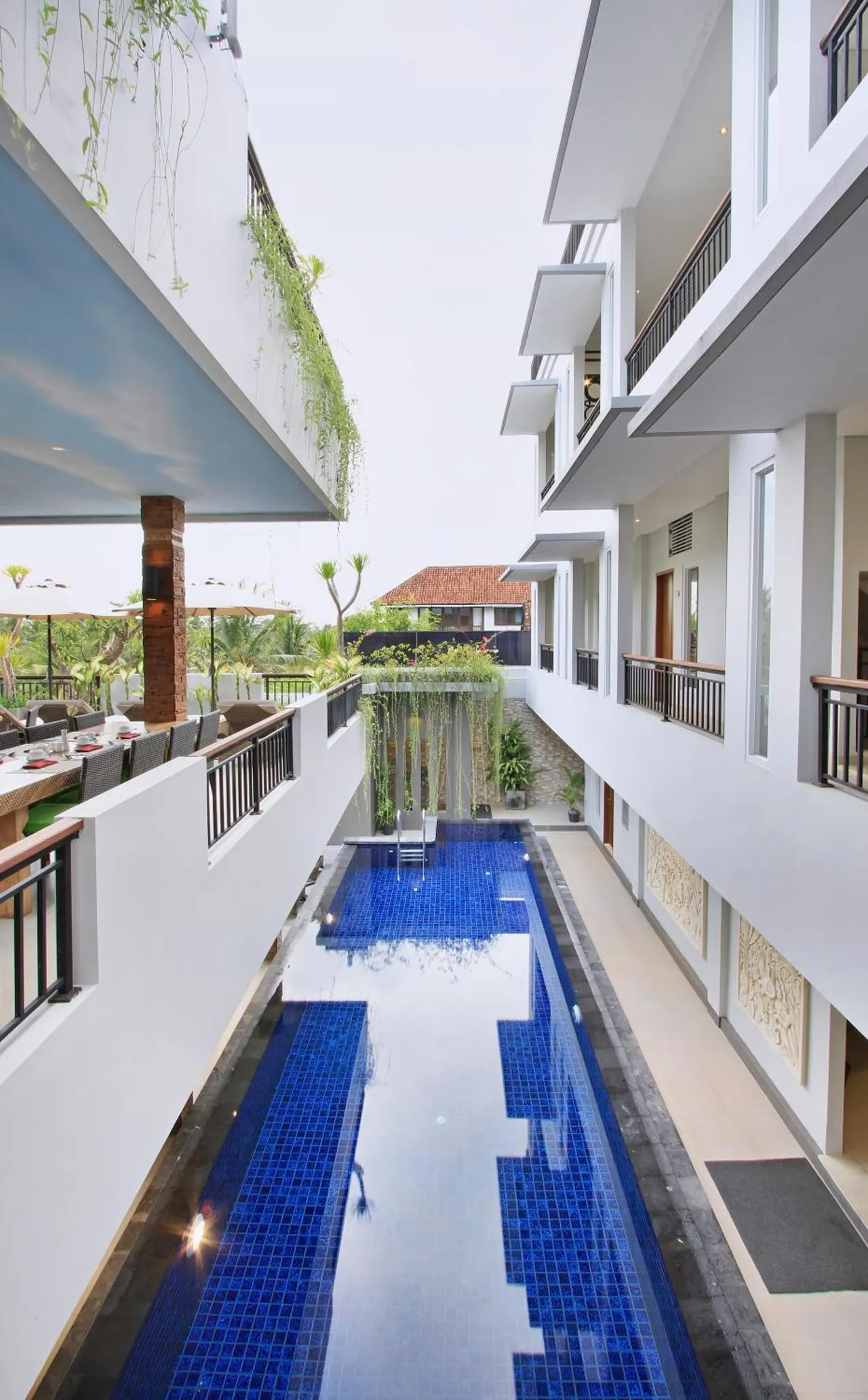 A long, narrow pool with blue mosaic tiles runs between two white buildings with balconies, and an outdoor dining area with umbrellas is visible on the left.