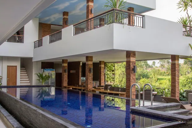 A long blue-tiled pool sits beneath a covered upper floor supported by brick pillars, with dining tables along one side and greenery visible through the open edge.