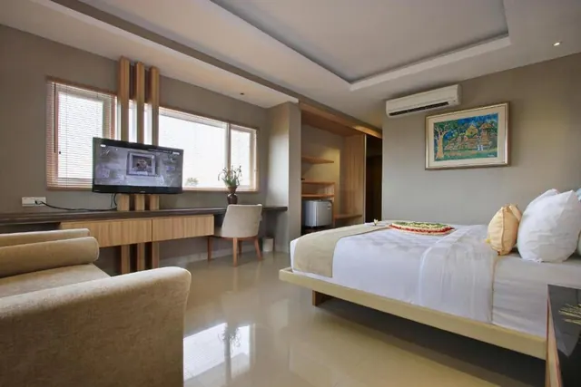A hotel bedroom with a made bed with white bedding and a flower garland, a wall-mounted television above a wooden desk with a chair, a sofa, a wall-mounted air conditioner, and a small fridge in a wooden alcove.