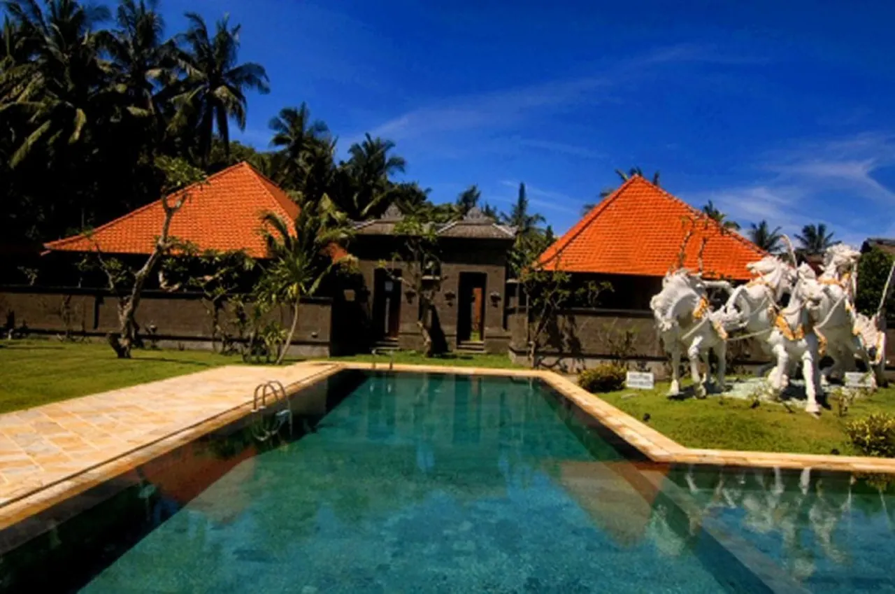 A rectangular swimming pool sits in front of Balinese-style buildings with orange tiled roofs, with white horse statues on the lawn to the right under a blue sky.