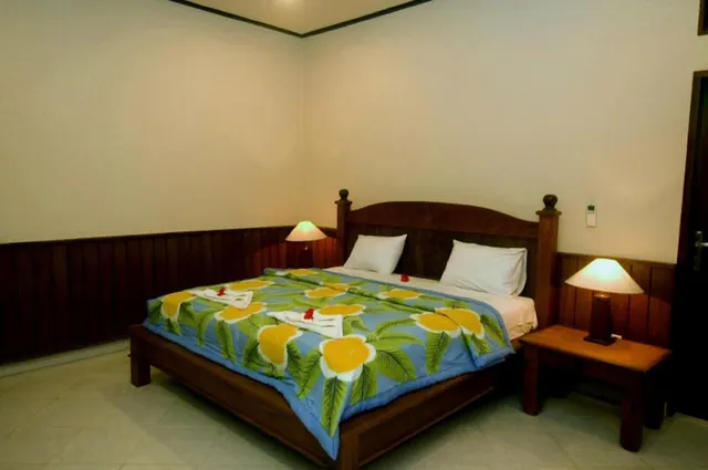 A bedroom with a wooden bed under a blue floral bedspread, two white pillows, folded towels decorated with red flowers, and two lit bedside lamps.