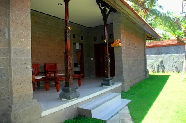 A covered veranda of a stone villa with carved wooden pillars and wooden armchairs with cushions, with steps leading down to a grassy lawn.