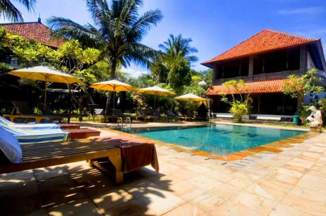 A rectangular swimming pool with a tiled surround, sun loungers and yellow umbrellas, with palm trees and a two-storey building with a red tiled roof behind.