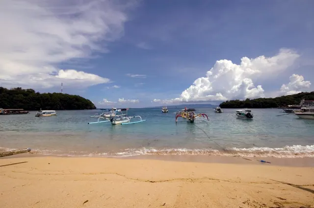 A sandy beach runs across the foreground to calm turquoise water where outrigger boats are anchored, with wooded headlands and cumulus clouds beyond.