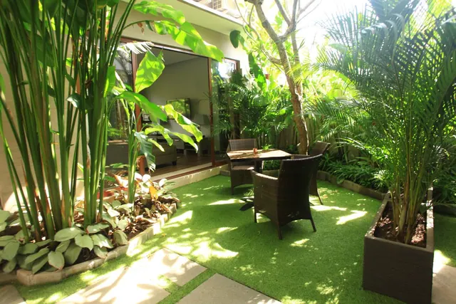 A private courtyard garden with artificial grass, a wicker dining set under a tree, tall tropical plants and planters, with glass doors to a room visible behind.