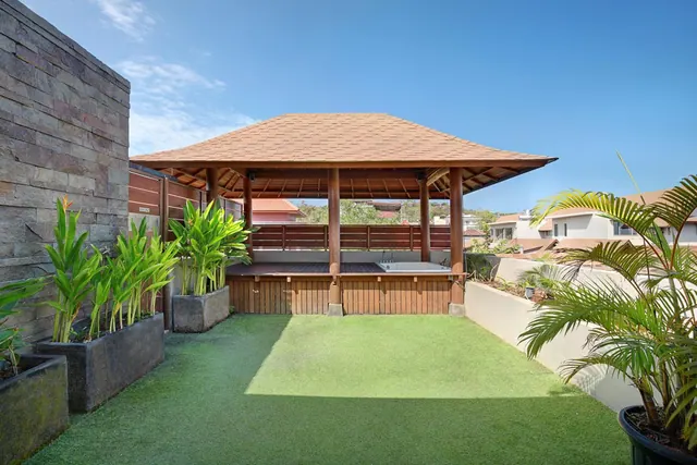 A wooden gazebo with a hot tub sits on an artificial grass terrace beside potted plants and a stone wall.