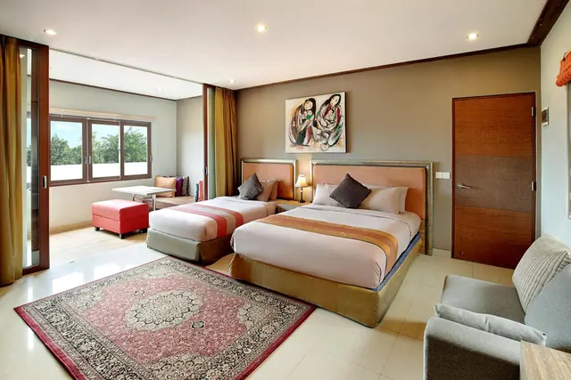 A bedroom with two large beds, a grey sofa, a patterned rug and a window looking onto trees.