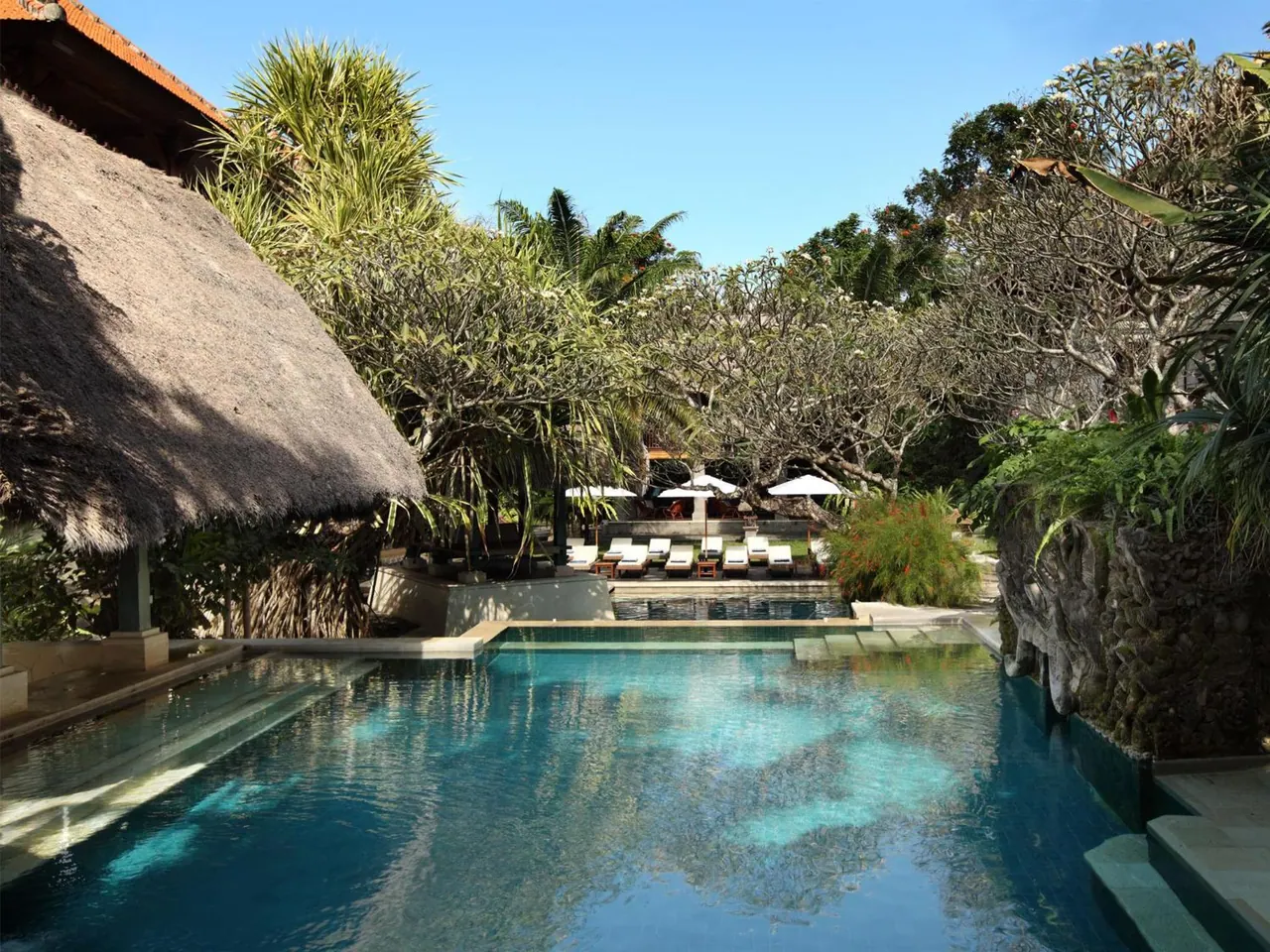 A long blue swimming pool with a thatched-roof pavilion on the left, tropical trees, and white sun loungers under umbrellas in the background.