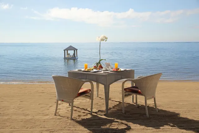 A breakfast table set with juice glasses, plates and a white orchid sits on a sandy beach facing the sea, with a small gazebo floating in the water.
