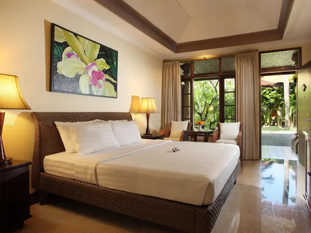 A bedroom with a large bed, two armchairs beside a small table, and glass doors opening onto a garden with tropical trees.