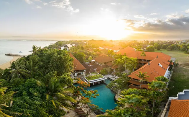 Aerial view of a resort with orange-tiled roofs, a turquoise pool, palm trees, and a sandy beach along the sea at sunset.
