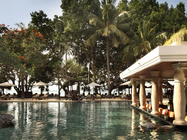 A large pool with sun loungers and umbrellas under palm trees, with a columned pavilion on the right and the sea visible beyond.