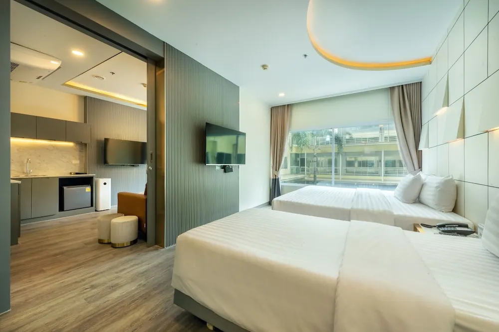 A hotel room with twin beds, a wall-mounted television, and a sliding doorway to a kitchenette and seating area, with a large window overlooking a pool.