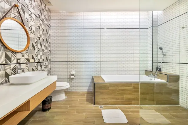 A bathroom with a patterned tile wall, a vessel sink on a wood and white vanity, a toilet, and a bathtub beside a glass shower screen with a handheld shower.