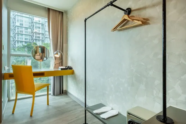 A guest room with a yellow desk and chair beside a window, a clothes rail with a wooden hanger, and slippers on a shelf.