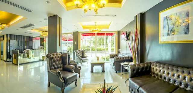 A lobby lounge with tufted leather armchairs and sofas around a coffee table, gold coffered ceilings with chandeliers, a reception desk on the left, and glass doors looking out onto greenery.