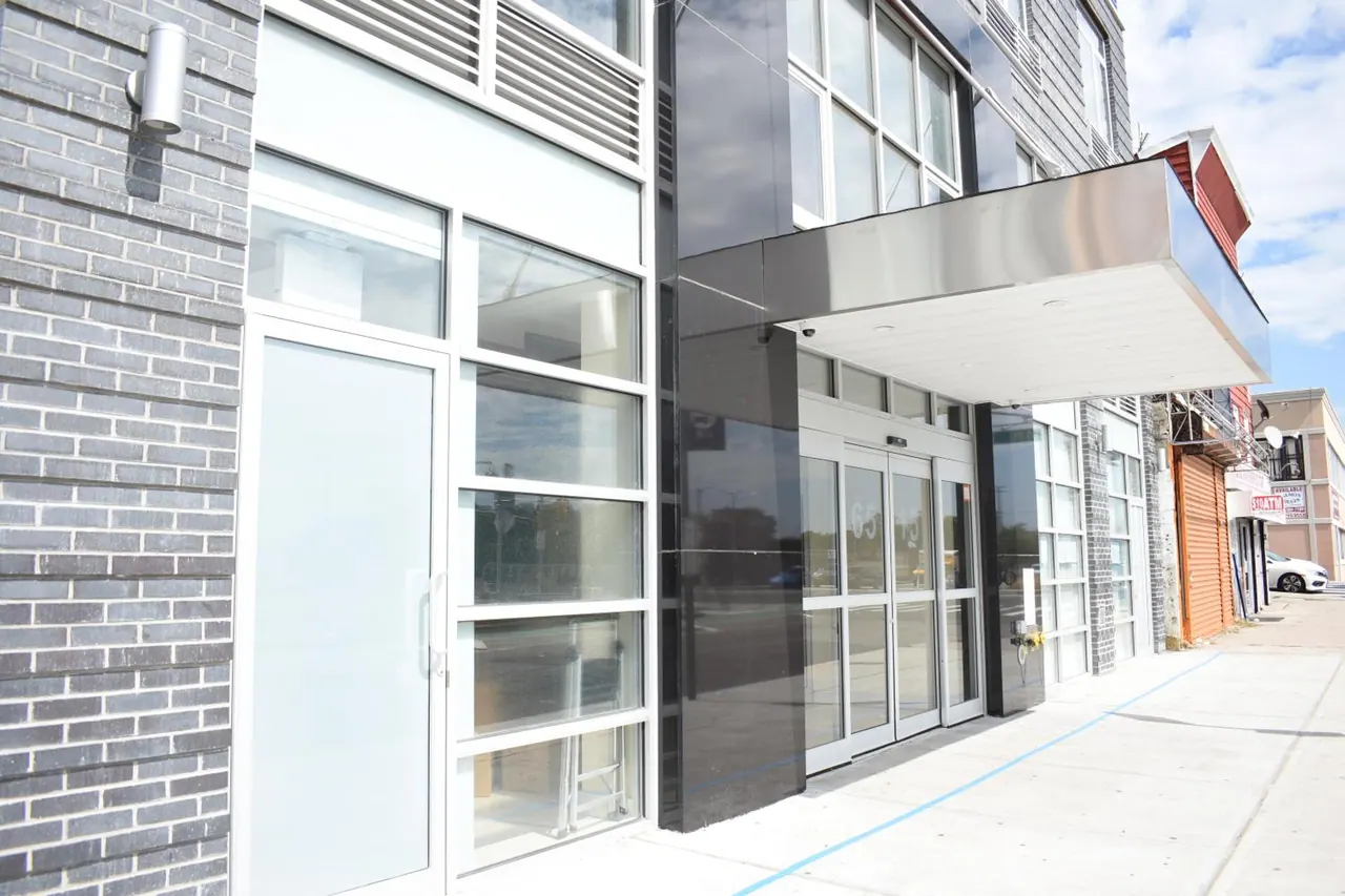 Glass entrance doors under a metal canopy on a building facade of grey brick and dark panels, with the sidewalk and street in front.