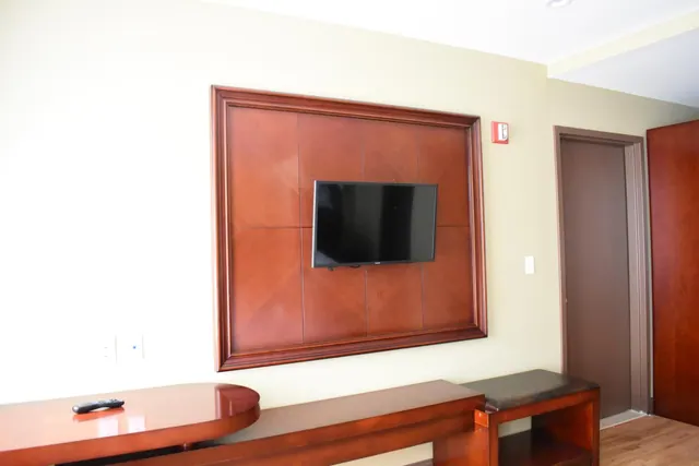 A wall-mounted flat-screen television sits inside a wood-panelled frame in a hotel room, with a remote control on a wooden desk beside it.