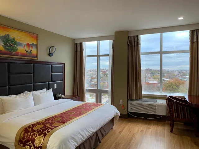 A bedroom with a king bed under a dark tufted headboard, a painting on the wall, a work desk and chair, and large windows looking over a city neighbourhood.