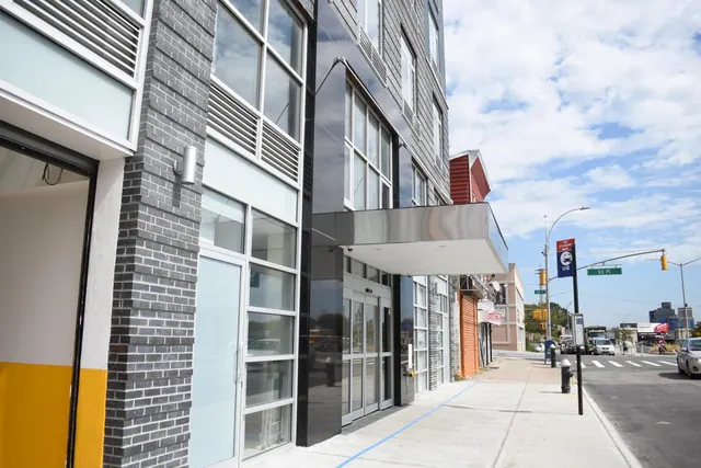 A modern building facade with dark brick and panel cladding, a glass entrance canopy and revolving doors, beside a street with parked cars and a traffic light.