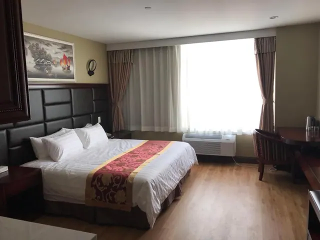 A hotel bedroom with a double bed under white bedding and a red and gold runner, a wall air conditioning unit under the window, and a wooden desk with a chair.