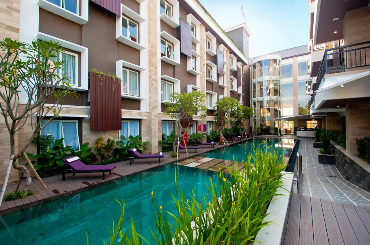 A long swimming pool in a hotel courtyard, with purple sun loungers on the wooden deck beside it and a multi-storey building behind.