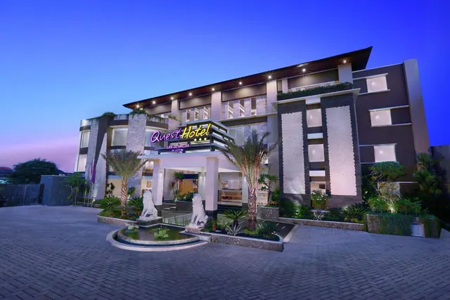 Exterior of the Quest Hotel Denpasar building at dusk, with an illuminated entrance, palm trees, lion statues and a circular fountain in the forecourt.