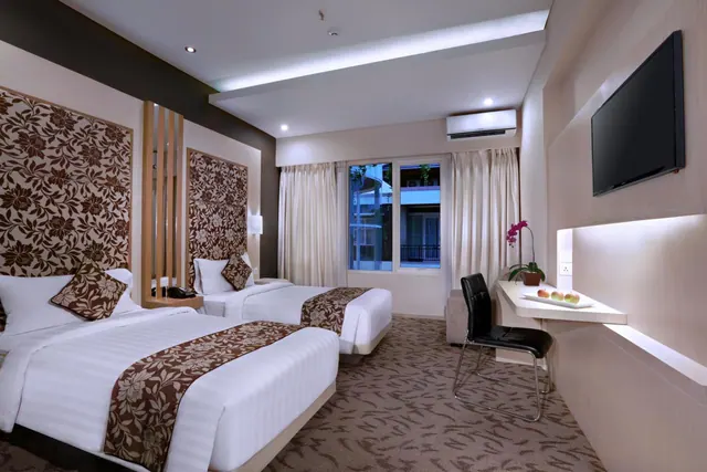 A hotel room with two beds with floral patterned headboards, a wall-mounted television, a desk with a chair, and a window looking out onto a neighbouring building.