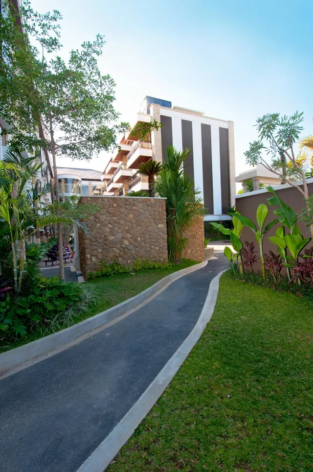A curved paved path runs through a lawn past a stone wall, tropical plants and a striped multi-storey building.