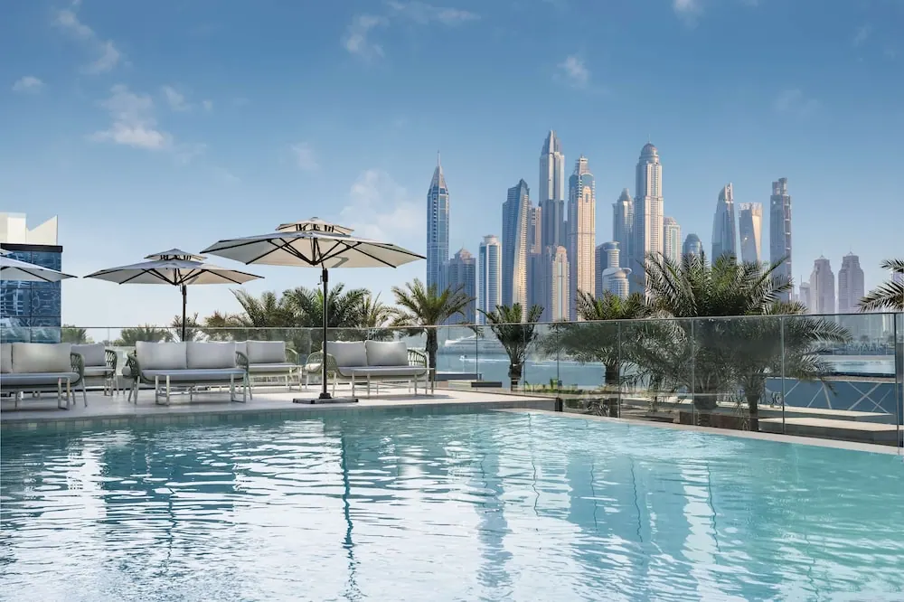 A rooftop swimming pool with white outdoor sofas and parasols, with a skyline of high-rise towers and palm trees behind it.