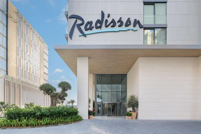 The entrance of a Radisson hotel shows its name sign on the white facade, a covered canopy over a glass revolving door, and landscaped planting along the paved driveway.