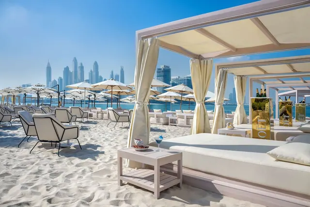 White daybeds and lounge chairs under cream canopies sit on a sandy beach, with parasols, the sea and a city skyline in the background.