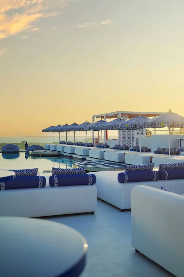 A poolside lounge at sunset with white sofas, blue cushions and patterned umbrellas beside a pool, with a sea horizon beyond the glass railing.
