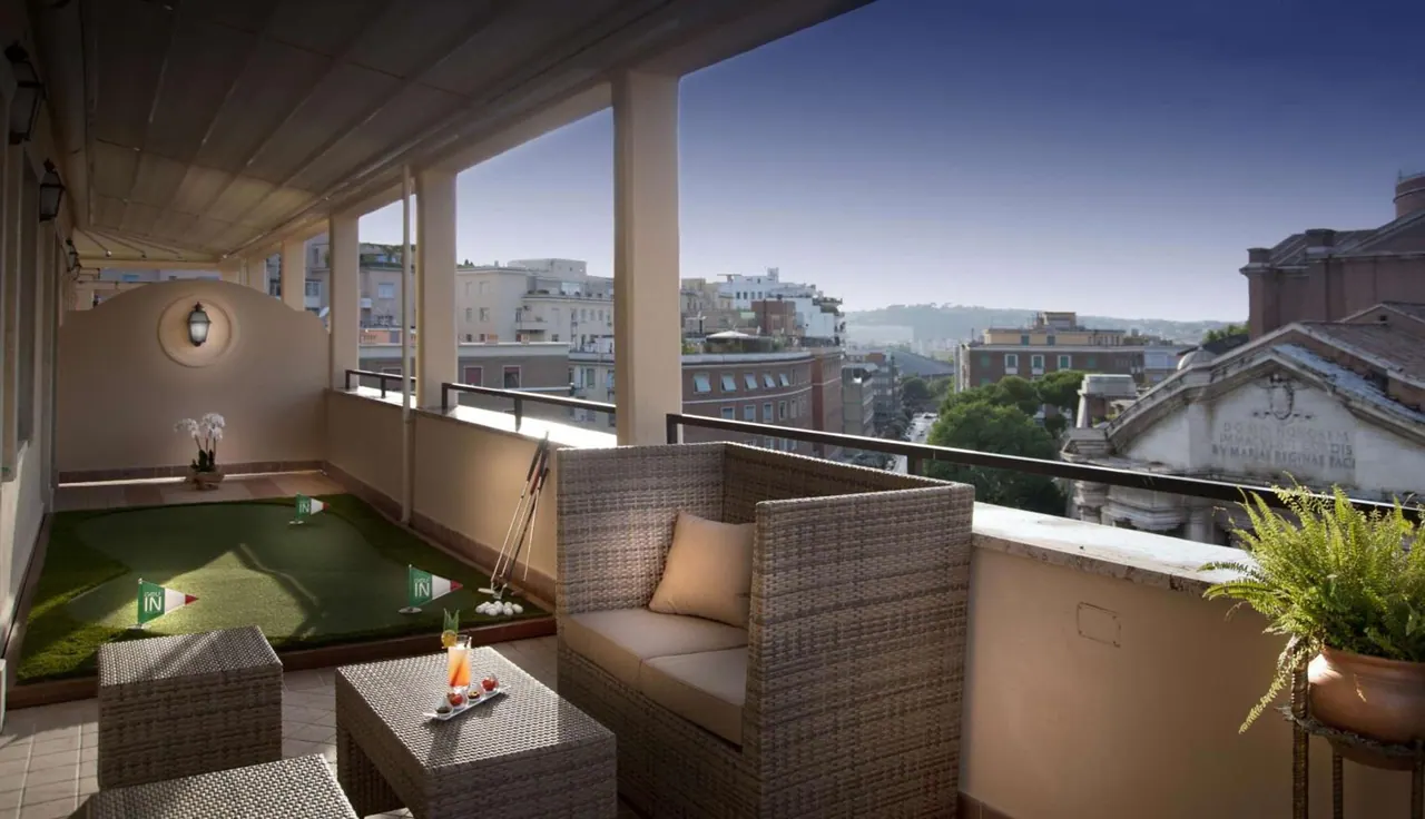 A covered rooftop terrace with a small putting green, a wicker sofa and coffee tables, overlooking city buildings and distant hills.