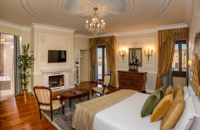 A large bedroom with a king bed, chandelier, fireplace, armchairs, wall-mounted television and an open balcony door, with a bathtub visible through the doorway at left.