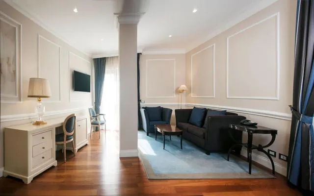 A staged suite living area with a dark grey sofa, armchair, blue rug, wall-mounted television and a white writing desk beside a central column.